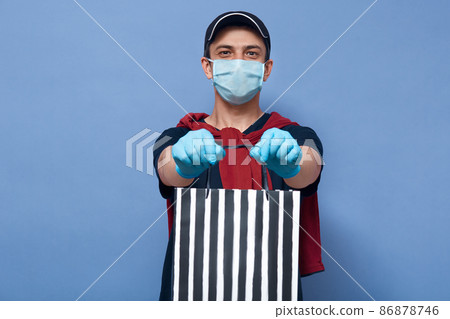 Closeup portrait of courier handing stripped shopping bag with online purchase, delivery man waring mask and gloves to prevent spreading dangerous corona virus all over world. Covid 19 concept. Closeup portrait of courier handing stripped shopping bag with online purchase, delivery man waring mask and gloves to prevent spreading dangerous corona virus all over world. Covid 19 concept. 86878746
