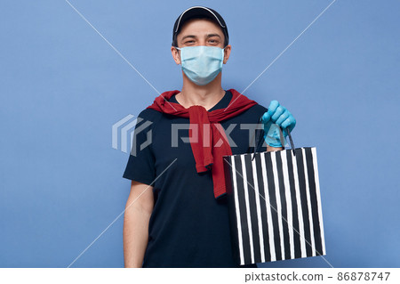 Closeup portrait of young handsome man working as courier, handing order in striped shopping bag, looking directly at camera, posing isolated over blue wall, delivers during coronavirus quarantine. 86878747