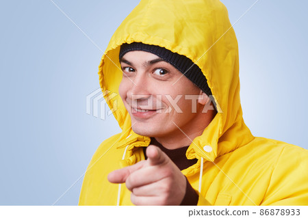 Happy man with dark appealing eyes wears yellow coat with anorak, points with fore finger diractly into camera, chooses someone, isolated over blue background. Selective focus. Man indicates at you 86878933