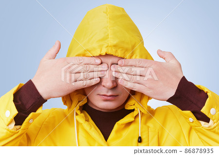 Serious young male wears yellow anorak and hood, covers eyes with hands, anticipates for surprise or tries to hide himself. Fashionable hipster guy gestures against light blue studio background 86878935