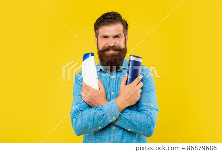 smiling brutal bearded man choosing hair conditioner bottle, presenting toiletries. 86879686