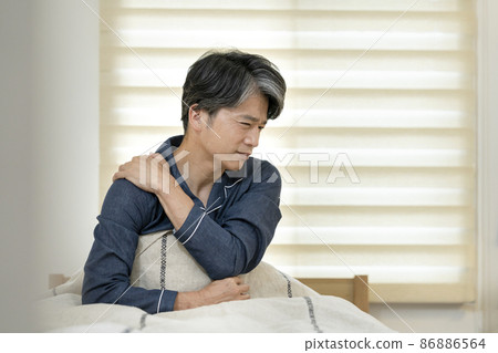 A man in his 50s sitting on the bed and holding his shoulders by hand 86886564