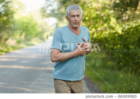 Photo of fatigue mature man feels pain in heart after jogging, has sullen expression, wears casual clothes, cares of his health. Handsome tired male has heart attack after active workout in open air 86889980