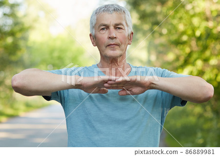 Confident grey haired male stretches hands, does physical exercise or workout in open air, has self assured facial expression stands outdoor, has healthy lifestyle. People, wellbeing and sport concept 86889981