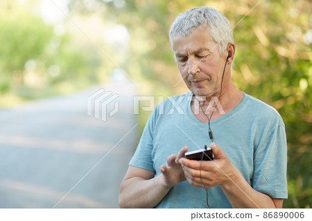 Serious elderly wrinkled male uses smart phone and earphones for listening music, focused into screen, has stroll in countryside, enjoys peaceful atmosphere, finds favourite melody in playlist Serious elderly wrinkled male uses smart phone and earphones for listening music, focused into screen, has stroll in countryside, enjoys peaceful atmosphere, finds favourite melody in playlist 86890006