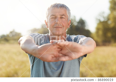 Elederly grey haired wrinkled male stretches hands, dressed in casual outfit, does sport outdoor on green field early in morning, enjoys sunrise and fresh air, has active lifestyle, likes sport Elederly grey haired wrinkled male stretches hands, dressed in casual outfit, does sport outdoor on green field early in morning, enjoys sunrise and fresh air, has active lifestyle, likes sport 86890019