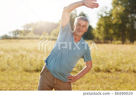 Outdoor shot of active senior male pensioner bends body, does physical exercises in coutryside, practices sport on old age, dressed in sports clothes. People, age and active lifestyle concept Outdoor shot of active senior male pensioner bends body, does physical exercises in coutryside, practices sport on old age, dressed in sports clothes. People, age and active lifestyle concept 86890020