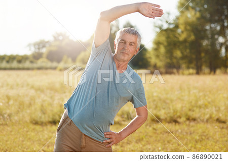 Athletic mature male with fit body, bends aside, does physical exercise outdoor, poses against green field background, breathes fresh air, has appealing look, wrinkled face, grey hair. Sport concept Athletic mature male with fit body, bends aside, does physical exercise outdoor, poses against green field background, breathes fresh air, has appealing look, wrinkled face, grey hair. Sport concept 86890021