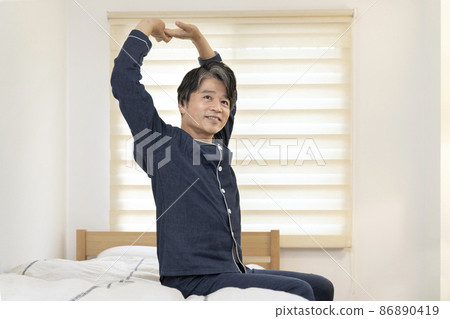A smiling man in his 50s in pajamas sitting on the bed and stretching A smiling man in his 50s in pajamas sitting on the bed and stretching 86890419