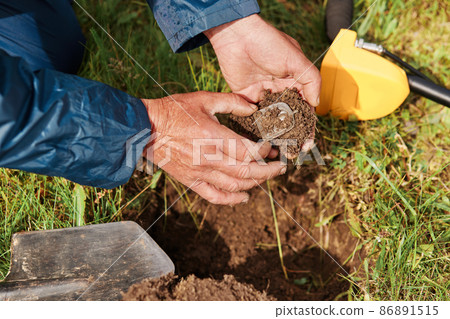 Unknown faceless treasure hunter holding his finding with ground in hands on background of grass, numismatist searching coins in field. Unknown faceless treasure hunter holding his finding with ground in hands on background of grass, numismatist searching coins in field. 86891515