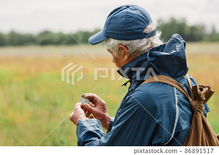 Gray haired man in jacket and cap treasure hunter looking at his finding, holding old coin under ground when searching in meadow or field. Gray haired man in jacket and cap treasure hunter looking at his finding, holding old coin under ground when searching in meadow or field. 86891517