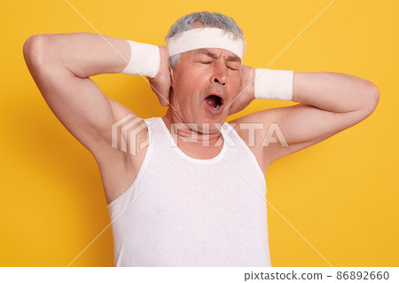Indoor shot of yawning senior man keeping hands up, keeps mouth opened and eyes closed, standing against yellow wall, looks sleepy, wearing white t shirt and headband. Indoor shot of yawning senior man keeping hands up, keeps mouth opened and eyes closed, standing against yellow wall, looks sleepy, wearing white t shirt and headband. 86892660