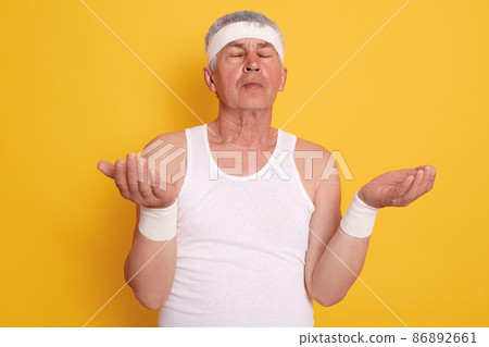 Closeup[ portrait of mature male with closed eyes, looks concentrated, posing with palms up, white haired man meditates at home while standing against yellow studio wall. Healthy lifestyle concept. Closeup[ portrait of mature male with closed eyes, looks concentrated, posing with palms up, white haired man meditates at home while standing against yellow studio wall. Healthy lifestyle concept. 86892661
