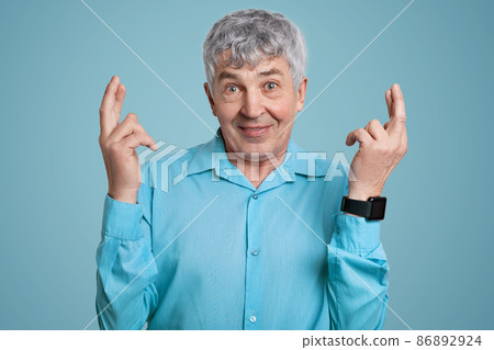 Horizontal shot of wrinkled man in blue formal shirt crosses fingers for good luck, being on pension, wears smartwatch, poses against blue background. Elderly people and body language concept 86892924