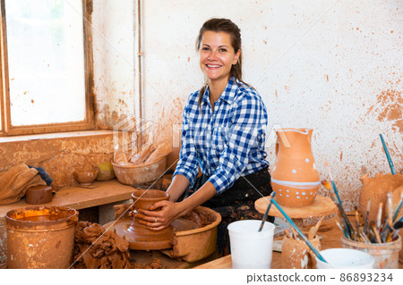 Female artisan in ceramics workshop with pottery wheel and various clay vessels 86893234