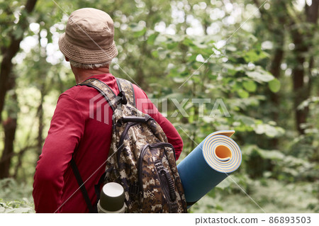 Senior backpackers walking along road in forest, eldery traveller wears red casual sweater, cap, rucksack, senior man keeps hands on hips, finds way where to go. Traveling and active holiday copncept. 86893503