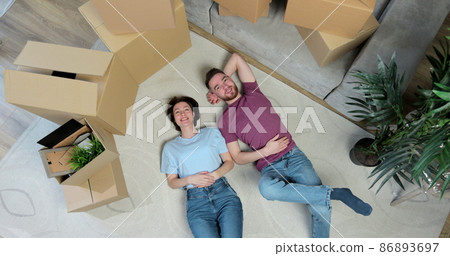 Happy but tired young couple lie on the floor of their new home. Young couple lying on floor of their new apartment in bedroom, talking and hugging. Carton boxes, carpet and bed are visible. Happy but tired young couple lie on the floor of their new home. Young couple lying on floor of their new apartment in bedroom, talking and hugging. Carton boxes, carpet and bed are visible. 86893697