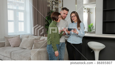 Couple standing in new house talking to woman real estate sales manager who is using tablet showing details. Buying home concept. Couple standing in new house talking to woman real estate sales manager who is using tablet showing details. Buying home concept. 86893807