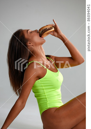 cheat meal concept. athlete woman is posing with fast food on light gray background, studio shot. young sporty woman is holding fast food close up. cheat meal concept. athlete woman is posing with fast food on light gray background, studio shot. young sporty woman is holding fast food close up. 86894041