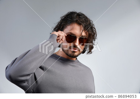 Fashion portrait of handsome elegant man with curly hair wearing gray sweater and glasses on gray background in studio 86894053
