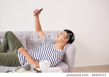 Handsome asian man relaxing with phone on the couch at home in the living room Handsome asian man relaxing with phone on the couch at home in the living room 86894315