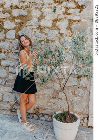 Girl Tourist Walking Through Ancient Narrow Street On A Beautiful Summer Day In MEDITERRANEAN MEDIEVAL CITY, OLD TOWN BUDVA, MONTENEGRO. Young Beautiful Cheerful Woman Walking On Old Street At 86895225