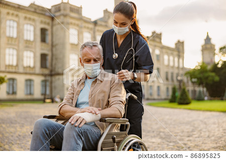 Young nurse in protective mask taking care of senior recovering patient in wheelchair with bandaged hand outdoors in the park near clinic 86895825