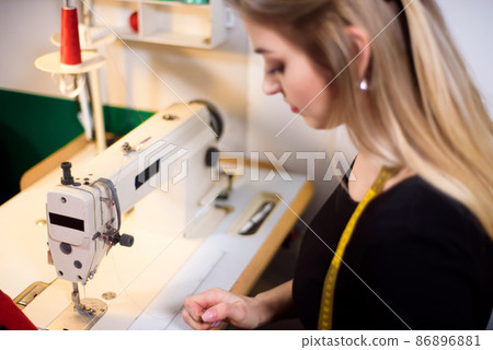 Tailor laboring at working place in atelier. Sewing machine, ruler, scissors, table lamp, threads on background. 86896881