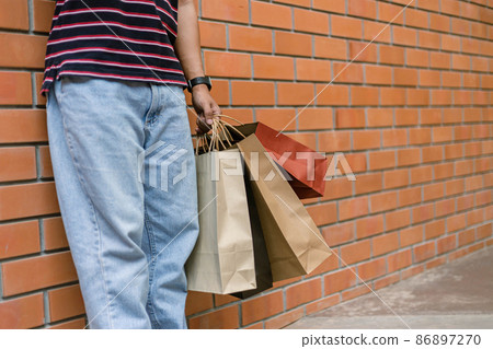shopping concept A person in light blue jeans and white shoes leaning on the wall of the building while holding a lot of paper shopping bags shopping concept A person in light blue jeans and white shoes leaning on the wall of the building while holding a lot of paper shopping bags 86897270