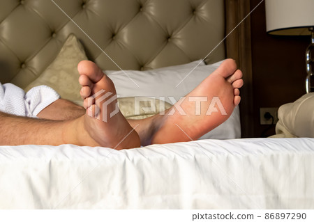Handsome male legs protrude from under a white blanket, in bed, in a comfortable apartment, in natural light. Copy space. Handsome male legs protrude from under a white blanket, in bed, in a comfortable apartment, in natural light. Copy space. 86897290