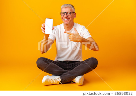 Senior handsome smiling man holding smartphone showing screen over yellow background 86898052