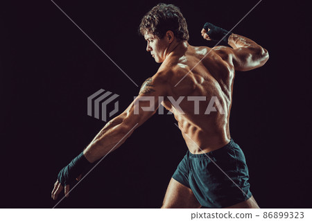 Side view of muscular shirtless boxer who strikes isolated on dark background. Sport concept Side view of muscular shirtless boxer who strikes isolated on dark background. Sport concept 86899323