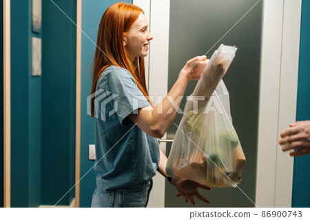 Side view of cheerful redhead female customer receiving groceries bag from unrecognizable delivery man on doorstep at apartment. Cropped shot of courier male delivering food meal to client at home. Side view of cheerful redhead female customer receiving groceries bag from unrecognizable delivery man on doorstep at apartment. Cropped shot of courier male delivering food meal to client at home. 86900743