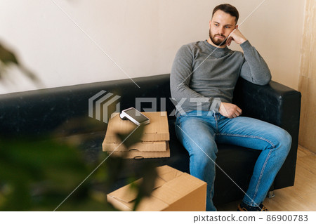 Bored courier male waiting customer while sitting on sofa with boxes pizza, cardboard box and payment POS wireless terminal in hall of apartment or office building. Concept of online shopping. 86900783