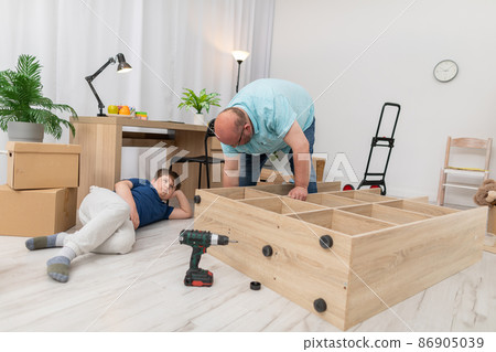 Dad laboriously assembles furniture and son just watches. Assembling furniture. 86905039