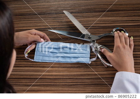 A young medical doctor is sitting at his desk and getting ready to cut the protective mask and thus end the ongoing pandemic and stringent restrictions on society. 86905222