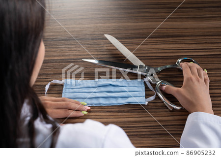A young medical doctor is sitting at his desk and getting ready to cut the protective mask and thus end the ongoing pandemic and stringent restrictions on society. A young medical doctor is sitting at his desk and getting ready to cut the protective mask and thus end the ongoing pandemic and stringent restrictions on society. 86905232