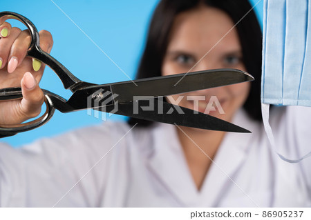 A young lady doctor is holding tailor's scissors in front of her with a protective mask ready for cutting. The end of the COVID-19 flu virus. A young lady doctor is holding tailor's scissors in front of her with a protective mask ready for cutting. The end of the COVID-19 flu virus. 86905237