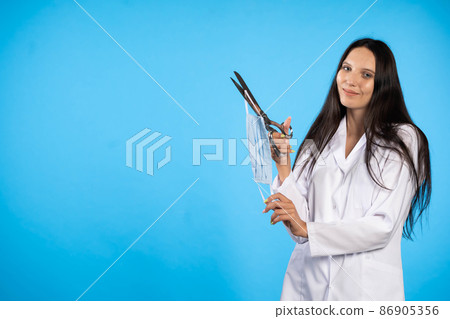 The doctor is holding large scissors and is going to cut the eraser of the protective masks. Global COVID-19 influenza virus pandemic. The doctor is holding large scissors and is going to cut the eraser of the protective masks. Global COVID-19 influenza virus pandemic. 86905356