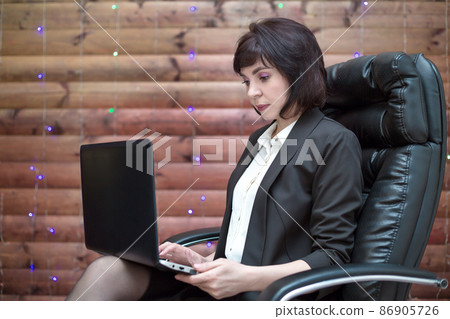 A brunette woman with a smile is sitting on a leather chair with a laptop in a formal suit in the office 86905726