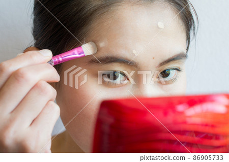 Portrait of Asian woman applying concealer on her facial skin with brush. Concealer is a type of cosmetic that is used to mask dark circles, age spots, large pores etc. Portrait of Asian woman applying concealer on her facial skin with brush. Concealer is a type of cosmetic that is used to mask dark circles, age spots, large pores etc. 86905733