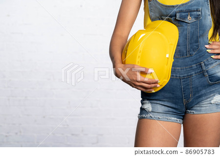 A woman holds a yellow construction protective helmet by her hand at the hip. Blue denim short pants called dungarees. A woman holds a yellow construction protective helmet by her hand at the hip. Blue denim short pants called dungarees. 86905783