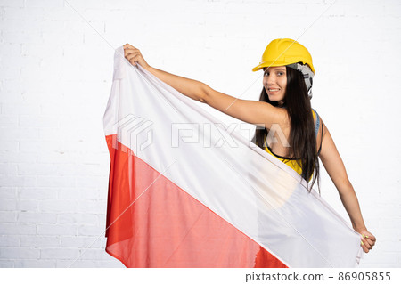 The construction worker is holding the national flag of Poland in her hands. The woman is wearing a helmet on her head. The construction worker is holding the national flag of Poland in her hands. The woman is wearing a helmet on her head. 86905855