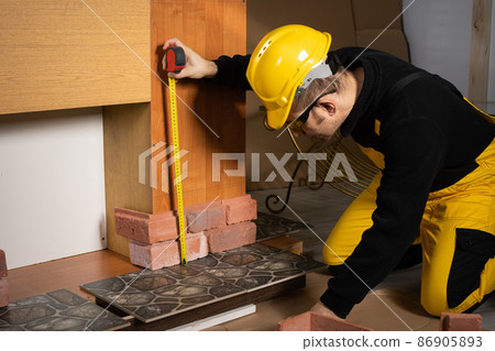 A construction worker in working clothes, kneeling, measures the retractable decorative tiles in the shape of old red brick with a tape measure. 86905893