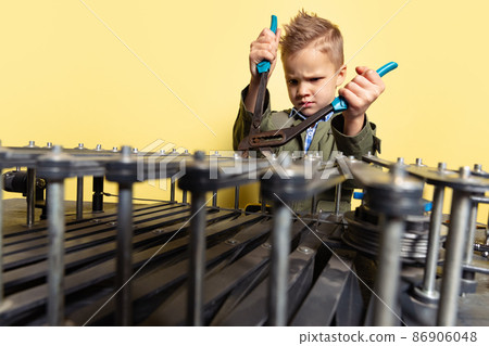 Looks serious. Little boy, child in image of auto mechanic or fitter in dungarees with work tools repairing auto motor isolated on yellow background. 86906048