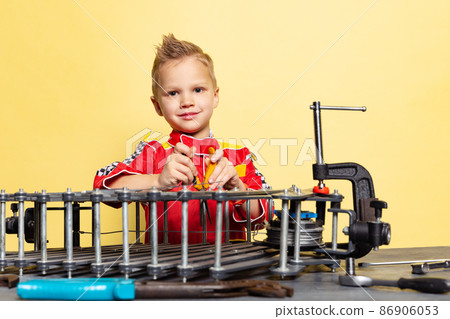 Cute little boy, child in image of auto mechanic or fitter in dungarees with work tools repairing auto motor isolated on yellow background. Cute little boy, child in image of auto mechanic or fitter in dungarees with work tools repairing auto motor isolated on yellow background. 86906053