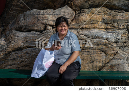 A middle-aged Asian woman looking at her telephone in front of a massive rock A middle-aged Asian woman looking at her telephone in front of a massive rock 86907008