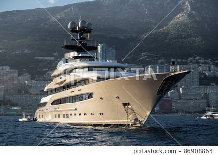 The huge yacht is beige color in sea at sunset, glossy board of the motor boat, the chrome plated handrail, megayacht is moored in sea, sun reflection on glossy board, mountains are on background 86908863