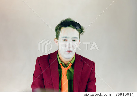 Person in bordo suit, shirt with yellow stripes tie looks down. Actor with white makeup on face, color eyes and green hair. Paint of clown or mime. Theater concept. Black glasses on head. Person in bordo suit, shirt with yellow stripes tie looks down. Actor with white makeup on face, color eyes and green hair. Paint of clown or mime. Theater concept. Black glasses on head. 86910014