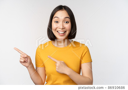 Portrait of smiling asian brunette girl in yellow tshirt, pointing fingers left, showing copy space, promo deal, demonstrating banner, standing over white background 86910686
